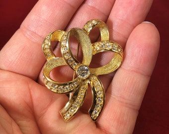 May include: A gold-tone brooch in the shape of a bow with clear rhinestones. The bow is made of metal and has a textured finish.