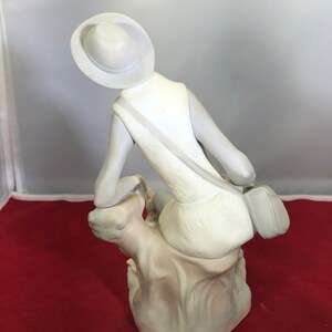 May include: White porcelain figurine of a person wearing a hat and sitting on a rock. The person is wearing a dress and a bag slung over their shoulder.