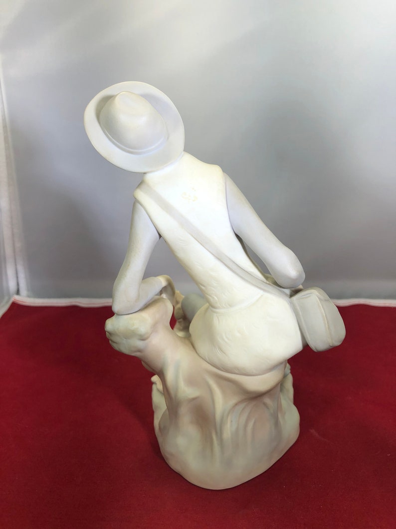 May include: White porcelain figurine of a person wearing a hat and sitting on a rock. The person is wearing a dress and a bag slung over their shoulder.