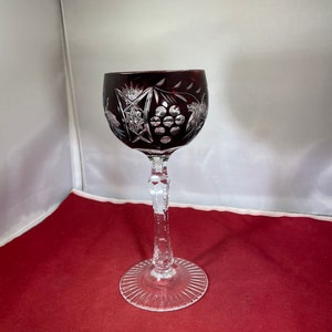 May include: A red crystal wine glass with a cut glass design of grapes and leaves. The glass has a long, clear stem and a clear, cut glass base.