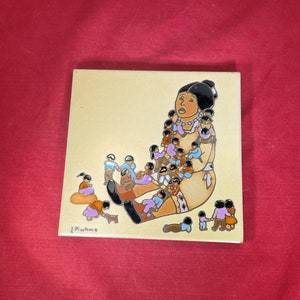 May include: A ceramic tile with a colorful illustration of a woman surrounded by many smaller figures. The woman is wearing a blue and white dress and has a necklace. The tile is signed "Muhme" in the bottom left corner.