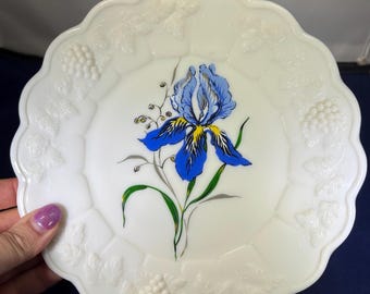 May include: White, scalloped-edge plate with embossed grape and leaf design around the rim. The center features a blue and yellow iris flower with green leaves. A decorative piece for serving or display.