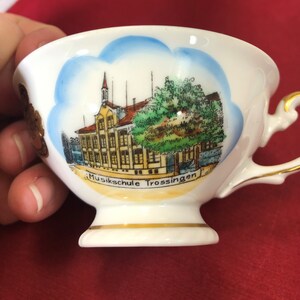 May include: A white porcelain teacup with a gold rim and a painted image of a building with the text "Musikschule Trossingen".