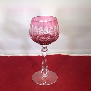 May include: A clear glass wine glass with a pink, diamond-shaped pattern. The glass has a long, slender stem and a round base.