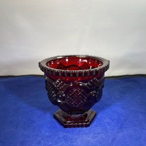 May include: A small, red glass bowl with a decorative pattern. The bowl has a scalloped rim and a pedestal base.