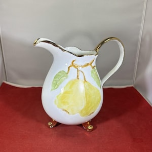 May include: White ceramic creamer with a gold rim and handle. The creamer features a hand-painted design of two yellow pears with green leaves. The creamer has three gold feet.