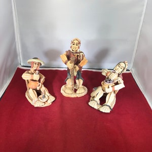 May include: Three hand-painted ceramic figurines depicting people in traditional clothing. The figures are seated and have detailed facial features. The figurines are brown, tan, and white.