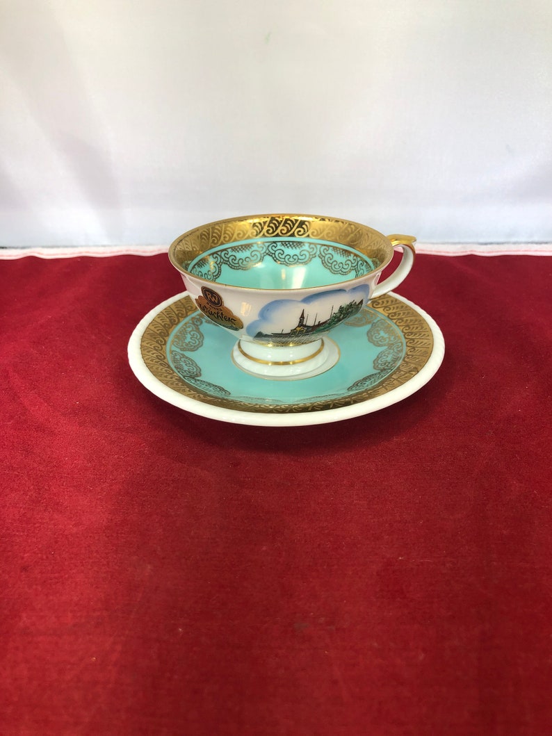 May include: A white teacup and saucer with a gold trim and a blue and white image of a cityscape. The teacup has a handle and the saucer has a scalloped edge.