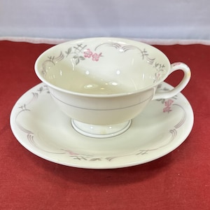 May include: A white teacup and saucer set with a delicate floral pattern in shades of pink and gray. The cup has a rounded shape and a handle, while the saucer has a scalloped edge.