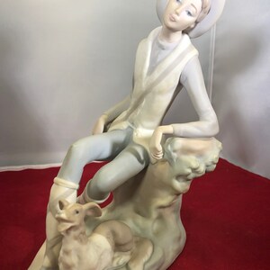 May include: A porcelain figurine of a young person wearing a hat and sitting on a rock with a dog at their feet. The figurine is painted in shades of blue, white, and brown.