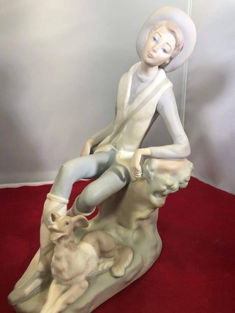 May include: A porcelain figurine of a young person wearing a hat and sitting on a rock with a dog at their feet. The figurine is painted in shades of blue, white, and brown.