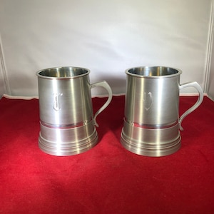 May include: Two silver pewter mugs with engraved initials, "J" and "V", on a red surface.