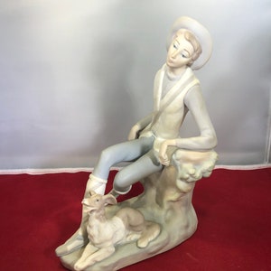 May include: A porcelain figurine of a young person sitting on a rock with a dog. The person is wearing a hat and a long-sleeved shirt. The dog is lying down and looking up at the person.