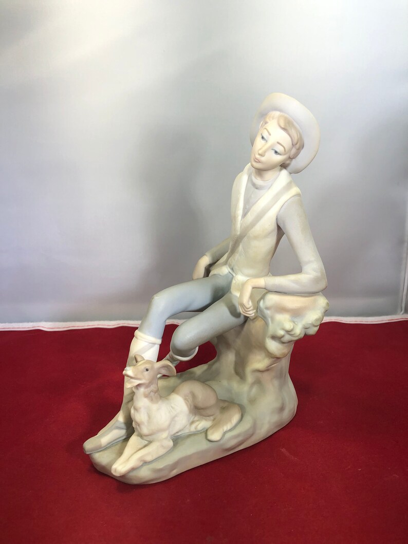 May include: A porcelain figurine of a young person sitting on a rock with a dog. The person is wearing a hat and a long-sleeved shirt. The dog is lying down and looking up at the person.