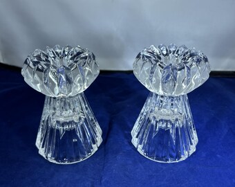 May include: Two clear glass candle holders. Each has a flower-shaped top and a flared base. The glass is faceted, creating a sparkling effect. The candle holders are displayed on a blue surface.