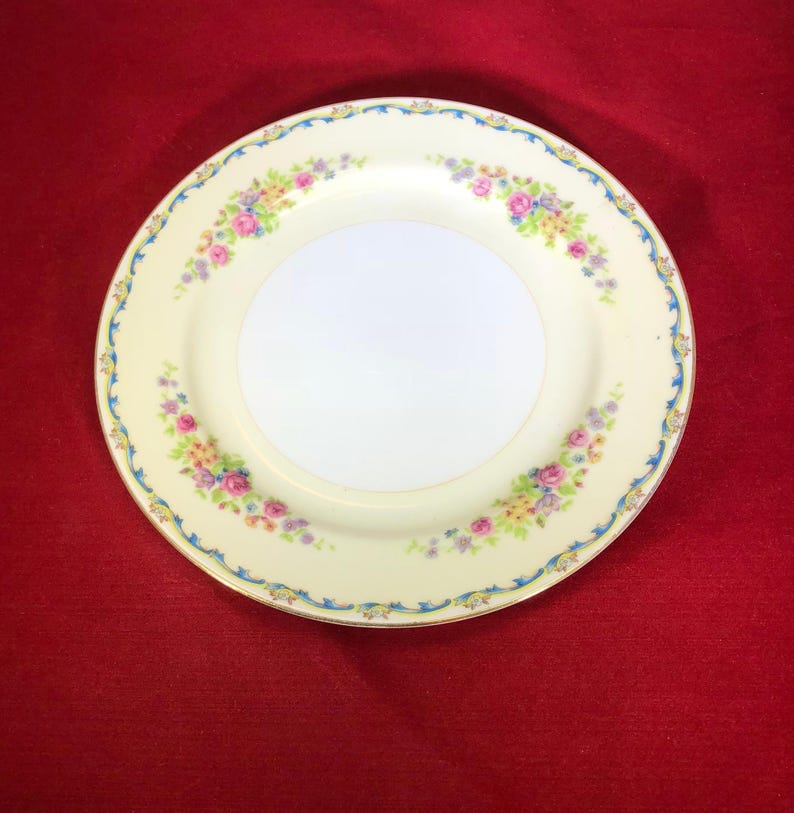 May include: A vintage floral design dinner plate with a cream colored background and a blue and gold trim. The plate features a delicate floral pattern with pink, yellow, and purple flowers.