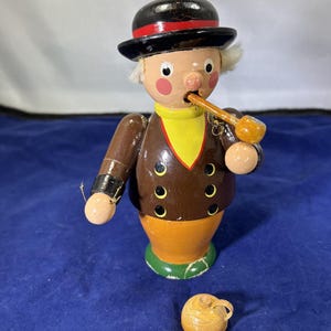 May include: A wooden figurine of a man wearing a black hat with a red band, brown jacket, and yellow collar. The figurine is holding a pipe and has a small, round, wooden jug next to it. The base is orange and green.