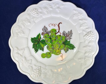 May include: White ceramic plate with a scalloped edge and embossed grape vine design. The center features a painted bunch of green grapes with green leaves and brown vines. A decorative piece for serving.
