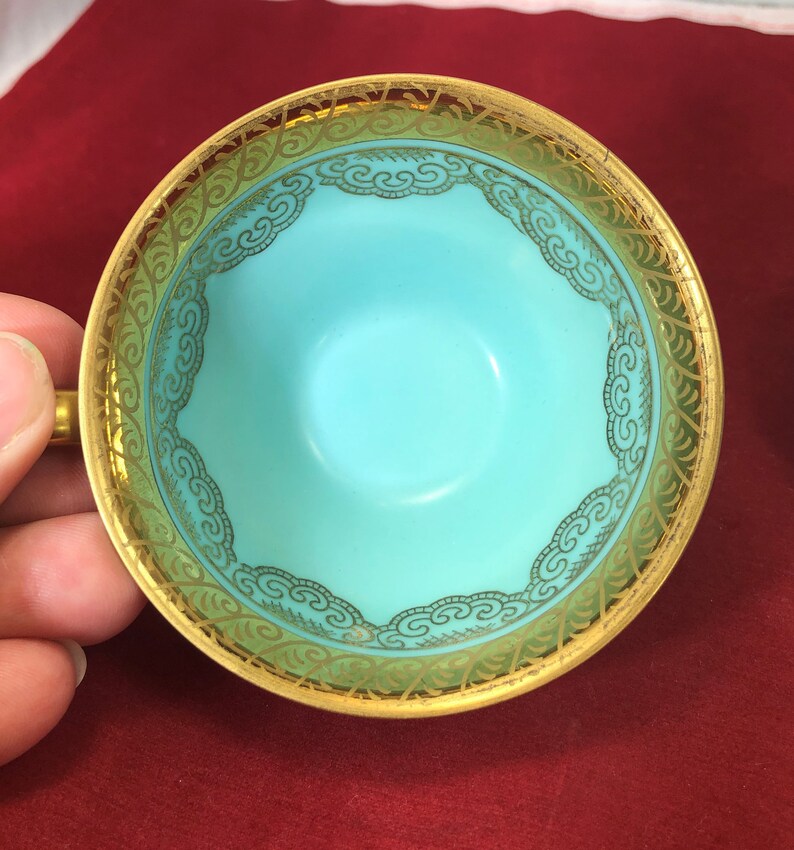 May include: A turquoise teacup with a gold rim and intricate gold design. The design features a repeating pattern of stylized clouds.