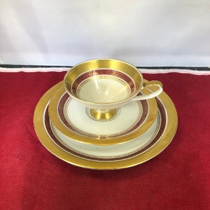 May include: A white teacup and saucer set with gold and burgundy trim. The cup and saucer have a delicate, ornate design.