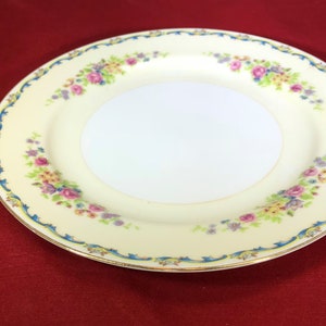 May include: A white porcelain plate with a floral design in shades of pink, yellow, and blue. The plate has a gold rim and a blue and gold border.