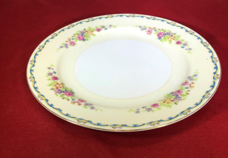 May include: A white porcelain plate with a floral design in shades of pink, yellow, and blue. The plate has a gold rim and a blue and gold border.