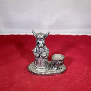 May include: A pewter figurine of a rabbit wearing a hat and holding a ladle, standing next to a small pewter pot with the year 1986 inscribed on it.