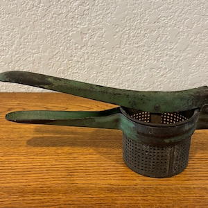 May include: A vintage green metal potato ricer with a handle. The ricer has a perforated metal base and is showing signs of wear.