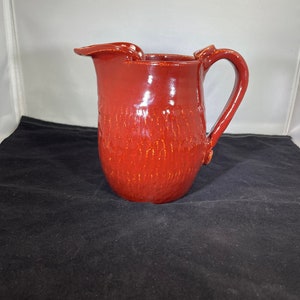 May include: A red ceramic pitcher with a textured surface and a wide, slightly curved spout. The pitcher has a handle on the side.