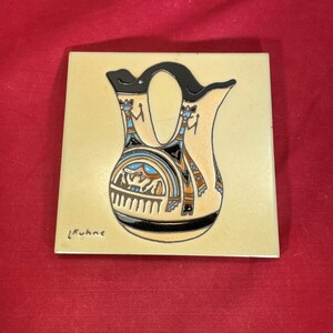 May include: A ceramic tile with a hand-painted design of a Native American pottery vase with black, brown, blue, and white accents. The tile is signed "L. Kuhne".