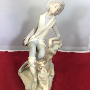 May include: A porcelain figurine of a young person wearing a hat and sitting on a rock with a dog. The figurine is painted in shades of white, gray, and brown.