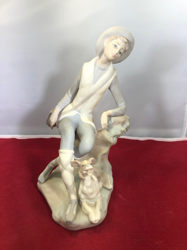 May include: A porcelain figurine of a young person wearing a hat and sitting on a rock with a dog. The figurine is painted in shades of white, gray, and brown.