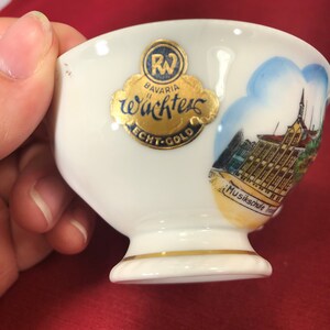 May include: White porcelain teacup with a gold rim and a gold sticker that says "Bavaria W&auml;chters Echt-Gold" and a picture of a building with the text "Musikschule Traunstein".