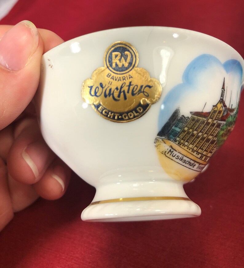 May include: White porcelain teacup with a gold rim and a gold sticker that says "Bavaria W&auml;chters Echt-Gold" and a picture of a building with the text "Musikschule Traunstein".