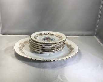May include: A set of six white porcelain dessert plates with gold trim and floral designs. The plates are stacked on a larger white porcelain plate with gold trim and a floral design.