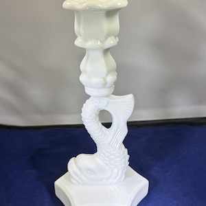May include: A white ceramic candlestick holder with a dolphin design. The candlestick features a square base, a dolphin body, and a fluted top for holding a candle. The dolphin's tail is visible. The candlestick is made of ceramic.