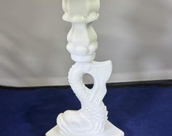 May include: A white ceramic candlestick holder with a dolphin design. The candlestick features a square base, a dolphin body, and a fluted top for holding a candle. The dolphin's tail is visible. The candlestick is made of ceramic.
