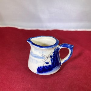 May include: A small white ceramic creamer with a blue and white windmill design. The creamer has a handle and a rounded body.