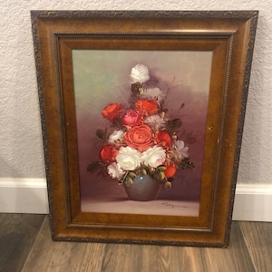 May include: A framed oil painting of a bouquet of red, white, and pink roses in a blue vase. The roses are arranged in a vase on a table with a light gray background.