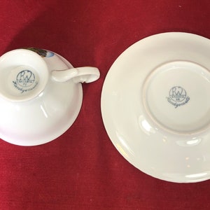 May include: A white porcelain teacup and saucer set with a blue and gold floral design. The bottom of the cup and saucer have a blue and white logo that reads "Bavaria RW Handgemalt".