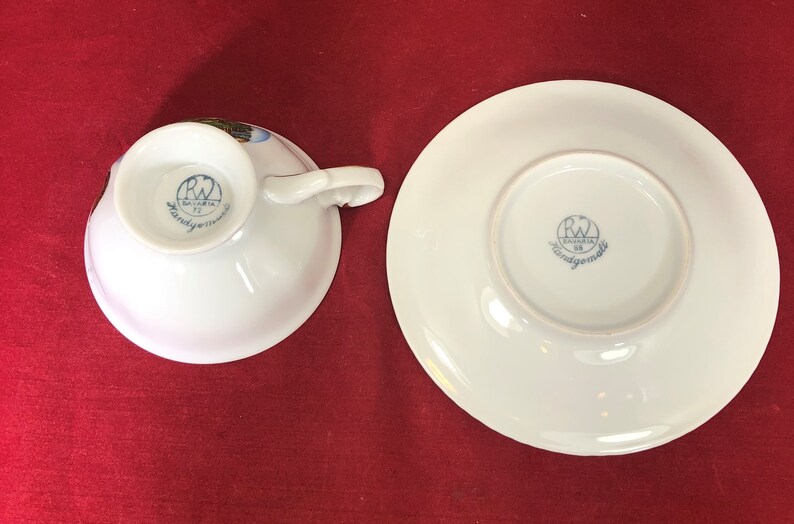 May include: A white porcelain teacup and saucer set with a blue and gold floral design. The bottom of the cup and saucer have a blue and white logo that reads "Bavaria RW Handgemalt".