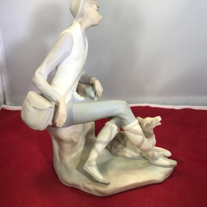 May include: A porcelain figurine of a person wearing a hat and sitting on a rock with a dog. The person is wearing a white vest and gray pants. The dog is brown and white.