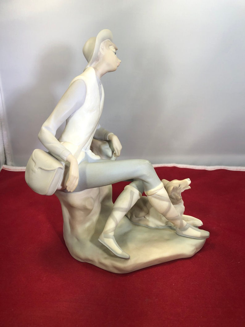 May include: A porcelain figurine of a person wearing a hat and sitting on a rock with a dog. The person is wearing a white vest and gray pants. The dog is brown and white.