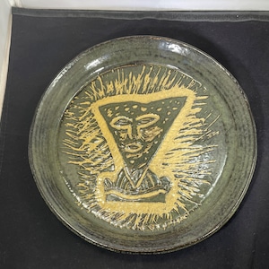 May include: A green ceramic plate with a raised, stylized face design in yellow glaze. The face is in a triangle shape with a crown-like design above it.