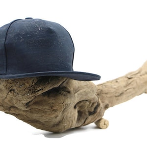 Könnte beinhalten: Eine dunkelblaue Baseballkappe mit einem gebogenen Schirm, die auf einem Stück Treibholz liegt.