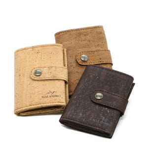 Puede incluir: Tres carteras de corcho en tonos marrones. Las carteras tienen una superficie texturizada y un cierre a presión. La marca "MALANDRO" es visible en una cartera. Las carteras están dispuestas sobre un fondo blanco.