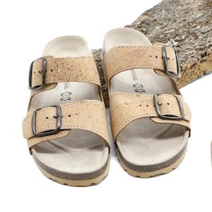 May include: A pair of light brown cork sandals with two adjustable straps and silver buckles. The sandals are displayed on a white surface with a piece of wood and a cork.