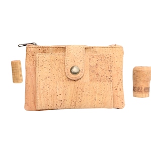 May include: A rectangular wallet made of light brown cork material with a speckled texture. The wallet features a top zipper closure and a front pocket secured by a flap with a metallic snap button. Two cylindrical cork stoppers are placed nearby.