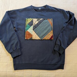 Peut inclure: Un sweat-shirt bleu marine avec un motif patchwork rectangulaire sur le devant. Le patchwork présente diverses pièces de tissu dans les tons de bleu, vert, marron et beige. Le sweat-shirt a des manches longues et un col rond.