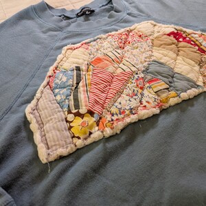 Peut inclure: Un sweat-shirt bleu avec un motif patchwork en forme d'éventail sur la poitrine. Le patchwork présente divers motifs et couleurs de tissu, notamment des textures florales, rayées et unies. Le sweat-shirt a un col rond et des manches longues.